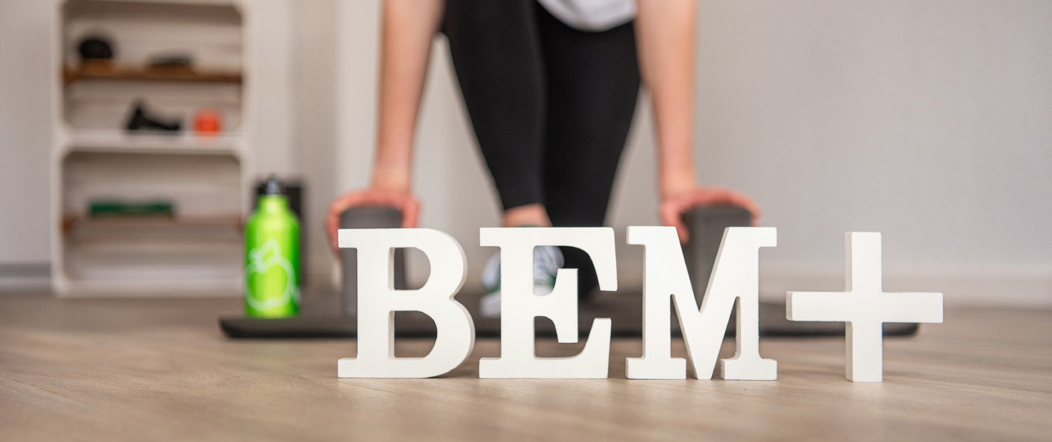 BEM+ Buchstaben im Fokus, zwei Yoga Klötze auf die sich eine Frau links und rechts mit den Händen stützt