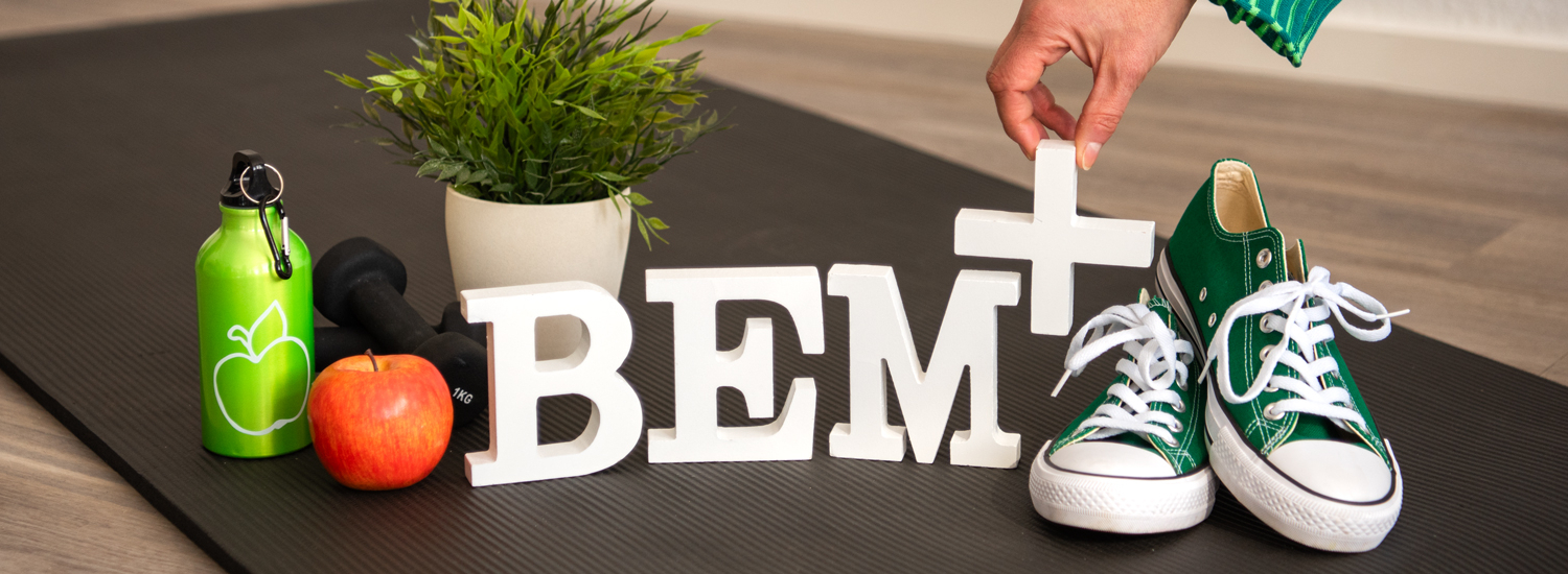 Header BEM+ Die Buchstaben BEM+ in weiß stehend auf einer schwarzen Yoga-Matte auf einem Holzfußboden, rechts daneben grüne Turnschuhe, links daneben ein roter Apfel sowie eine grüne Trinkflasche und schwarze Hanteln, im Hintergrund eine Grünpflanze