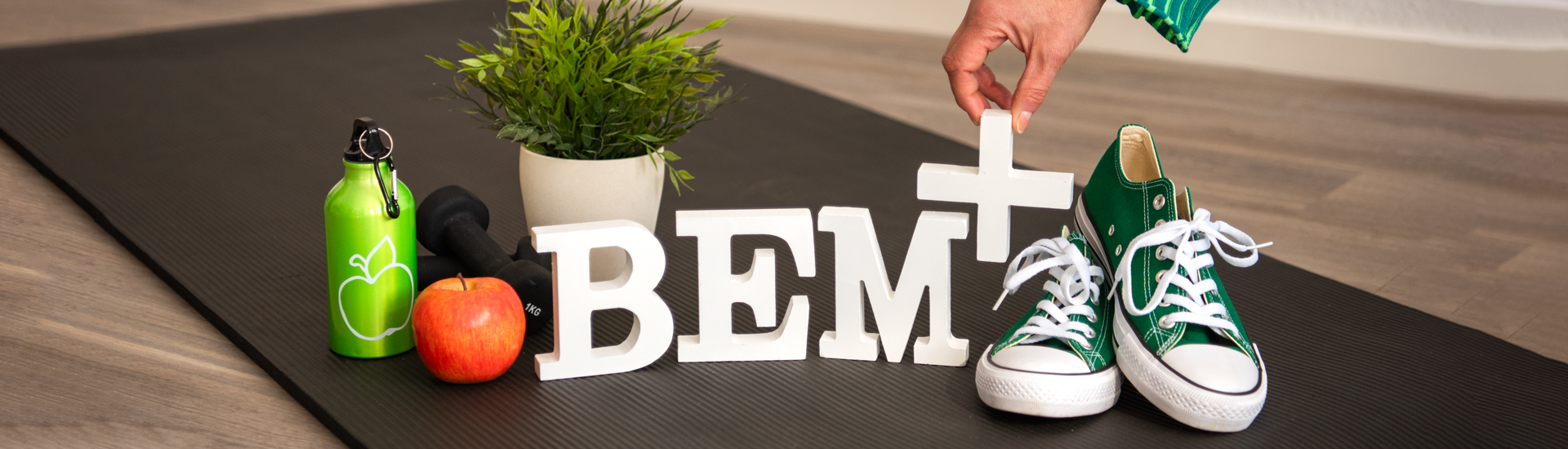 Die Buchstaben BEM+ in weiß stehend auf einer schwarzen Yoga-Matte auf einem Holzfußboden, rechts daneben grüne Turnschuhe, links daneben ein roter Apfel sowie eine grüne Trinkflasche und schwarze Hanteln, im Hintergrund eine Grünpflanze