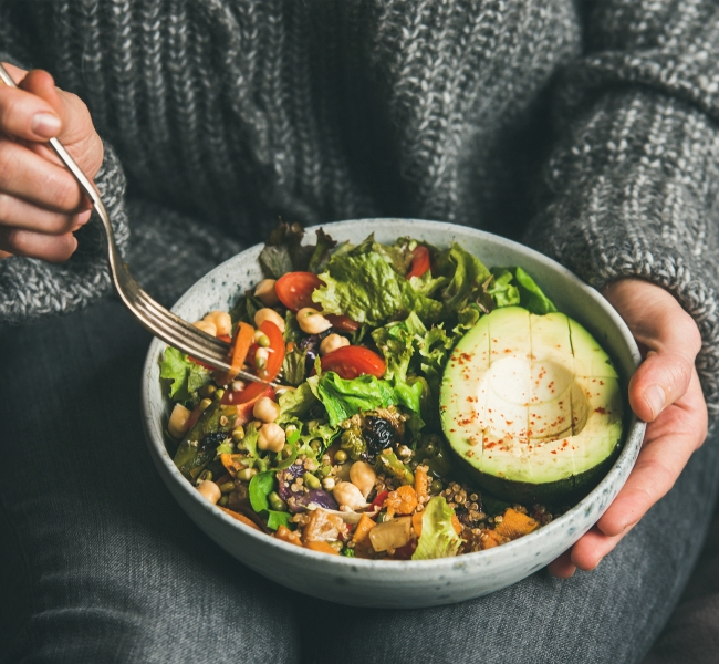 Bild Person in grauer Kleidung hat eine Bowl mit Salat und Avocado in der linken Hand und in der rechten Hand eine Gabel